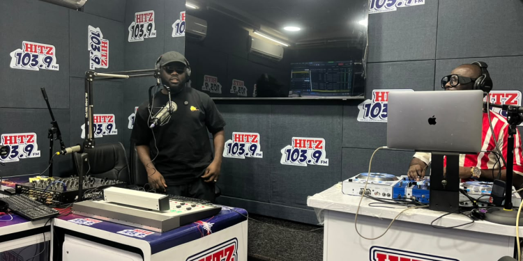 A guest in a black cap and T-shirt stands at the microphone in the Hitz FM studio as a presenter works the desk during the Multimedia Group and Influencer Africa takeover
